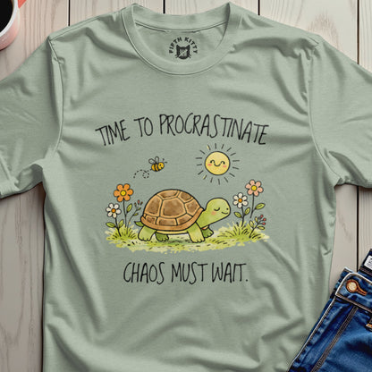 Green t-shirt with a cartoon turtle, bee, sun, and flowers, reading "TIME TO PROCRASTINATE CHAOS MUST WAIT."
