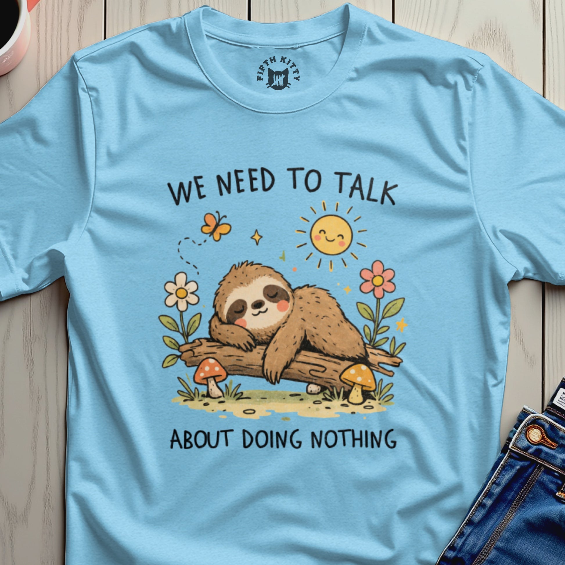 Light blue t-shirt with a sleeping sloth on a log, sun, flowers, and mushrooms.