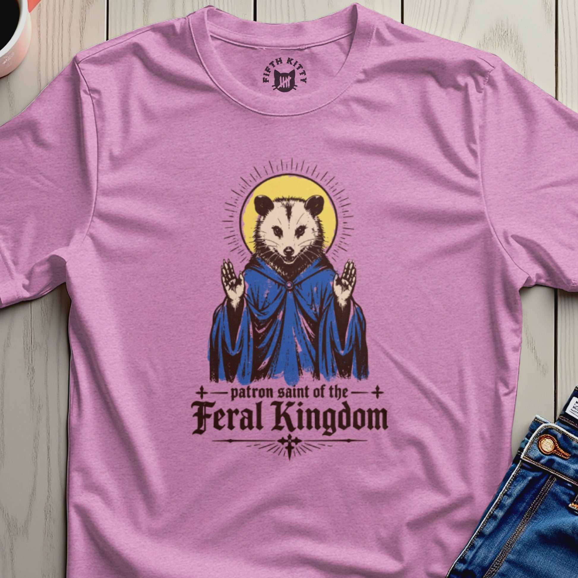 Pink t-shirt with a possum wearing a blue robe, halo, and text "patron saint of the FERAL KINGDOM".