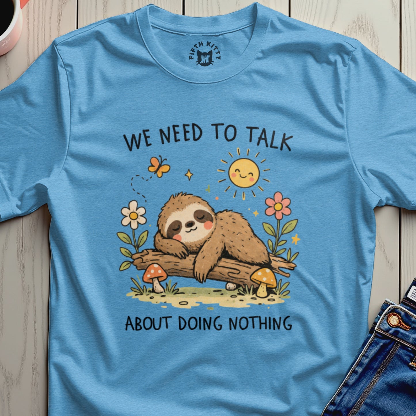 Blue t-shirt with a sleeping sloth on a log, surrounded by flowers and mushrooms.