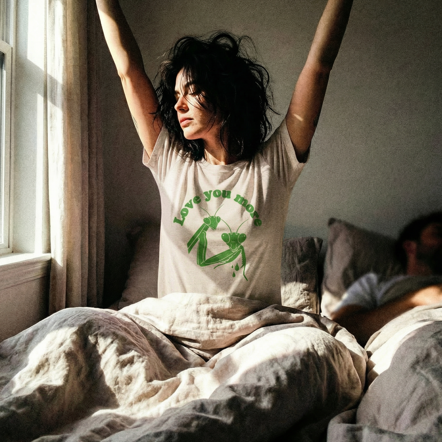 Person sitting on a bed with arms raised, wearing a t-shirt with green text and graphics. the text says "love you more" with a graphic of a praying mantis on it holding the head of it's lover
