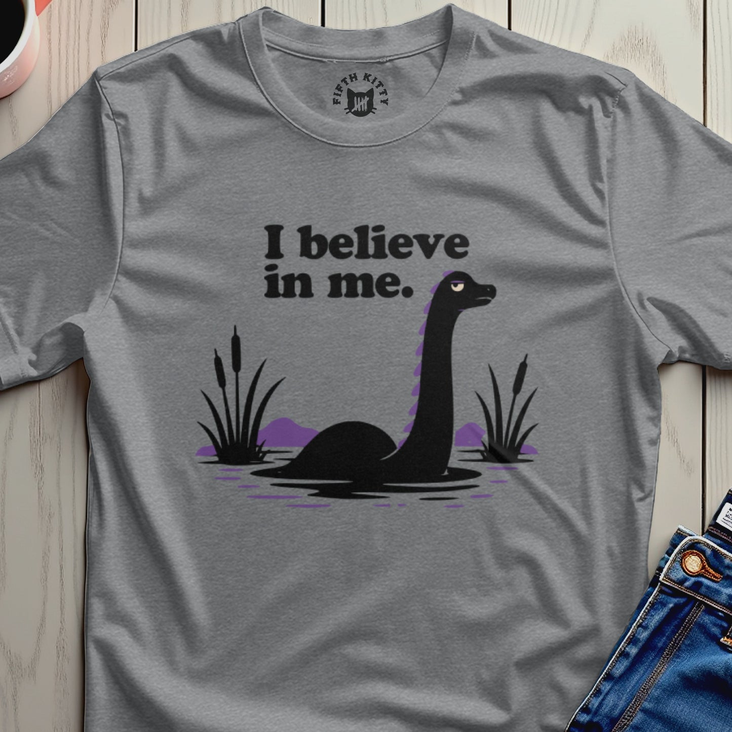 Gray t-shirt with black graphic of a Loch Ness monster and purple hills, reading "I believe in me."
