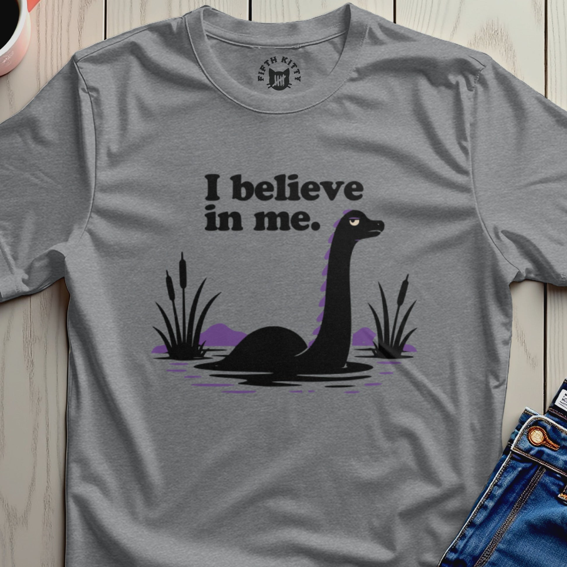 Gray t-shirt with black graphic of a Loch Ness monster and purple hills, reading "I believe in me."