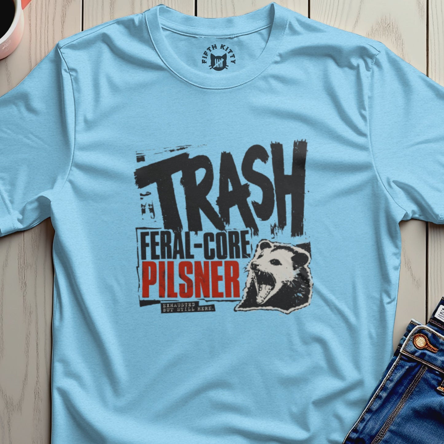 Light blue t-shirt with "TRASH FERAL-CORE PILSNER" text and raccoon graphic.
