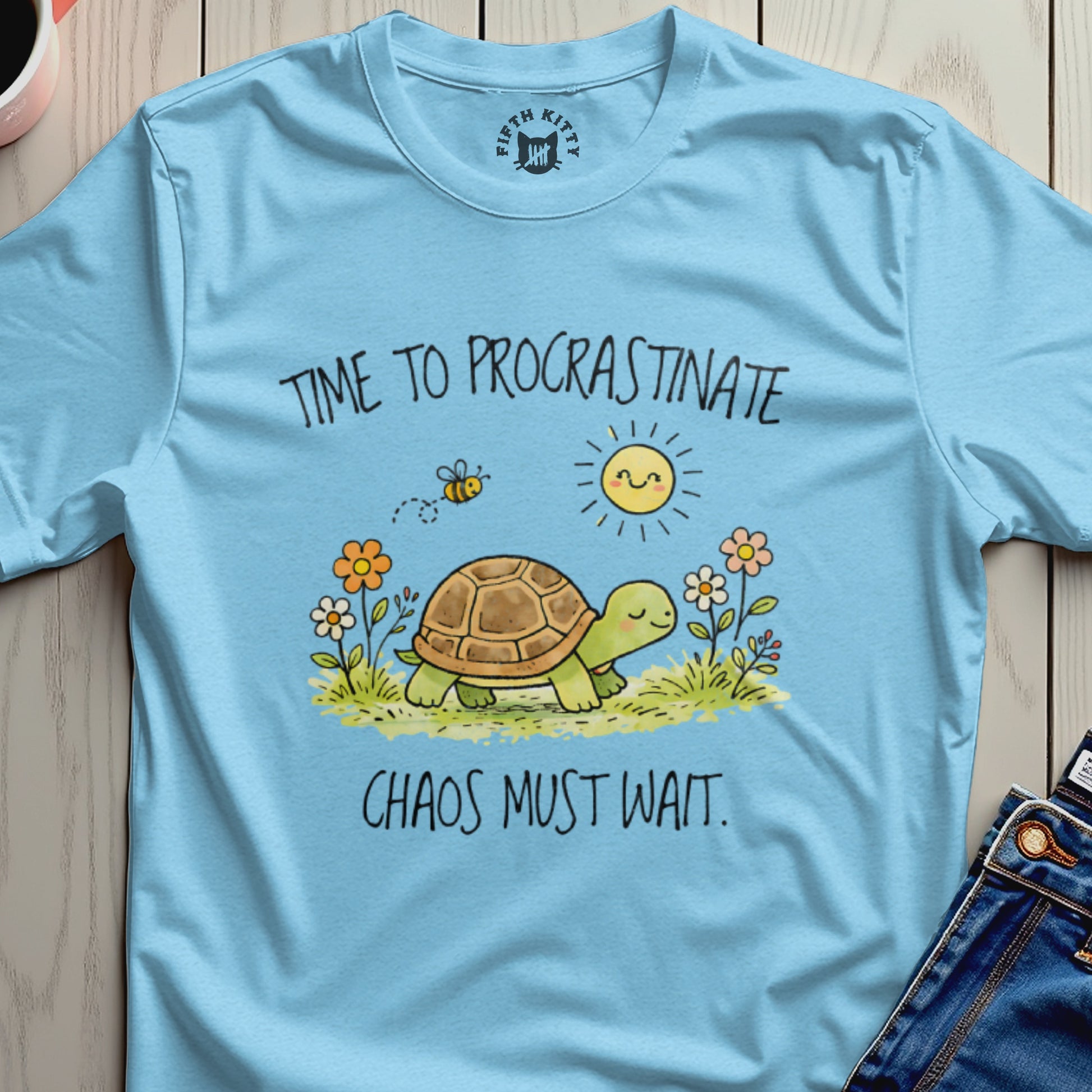 Light blue t-shirt with a turtle, bee, sun, and flowers, reads "TIME TO PROCRASTINATE CHAOS MUST WAIT."