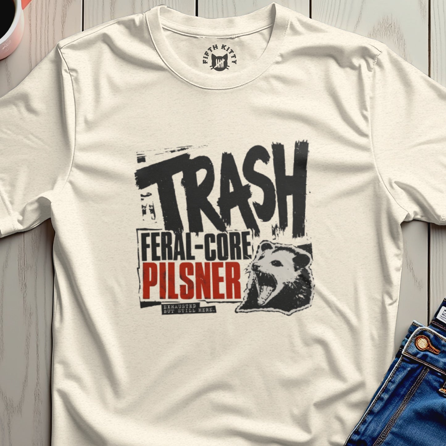 Cream t-shirt with black "TRASH FERAL-CORE PILSNER" text and raccoon graphic.