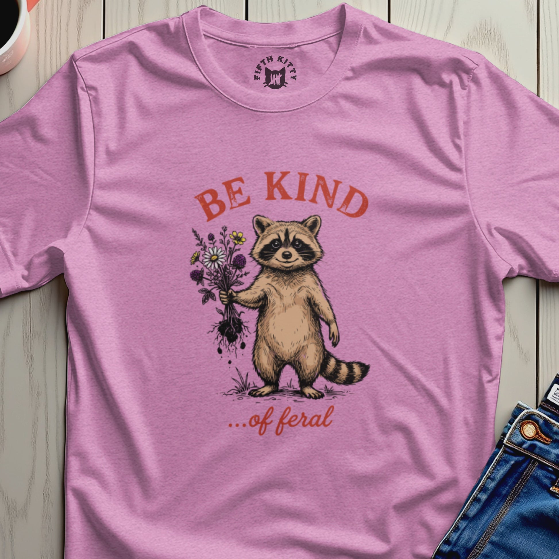 Pink t-shirt with raccoon holding flowers and text "BE KIND ...of feral".
